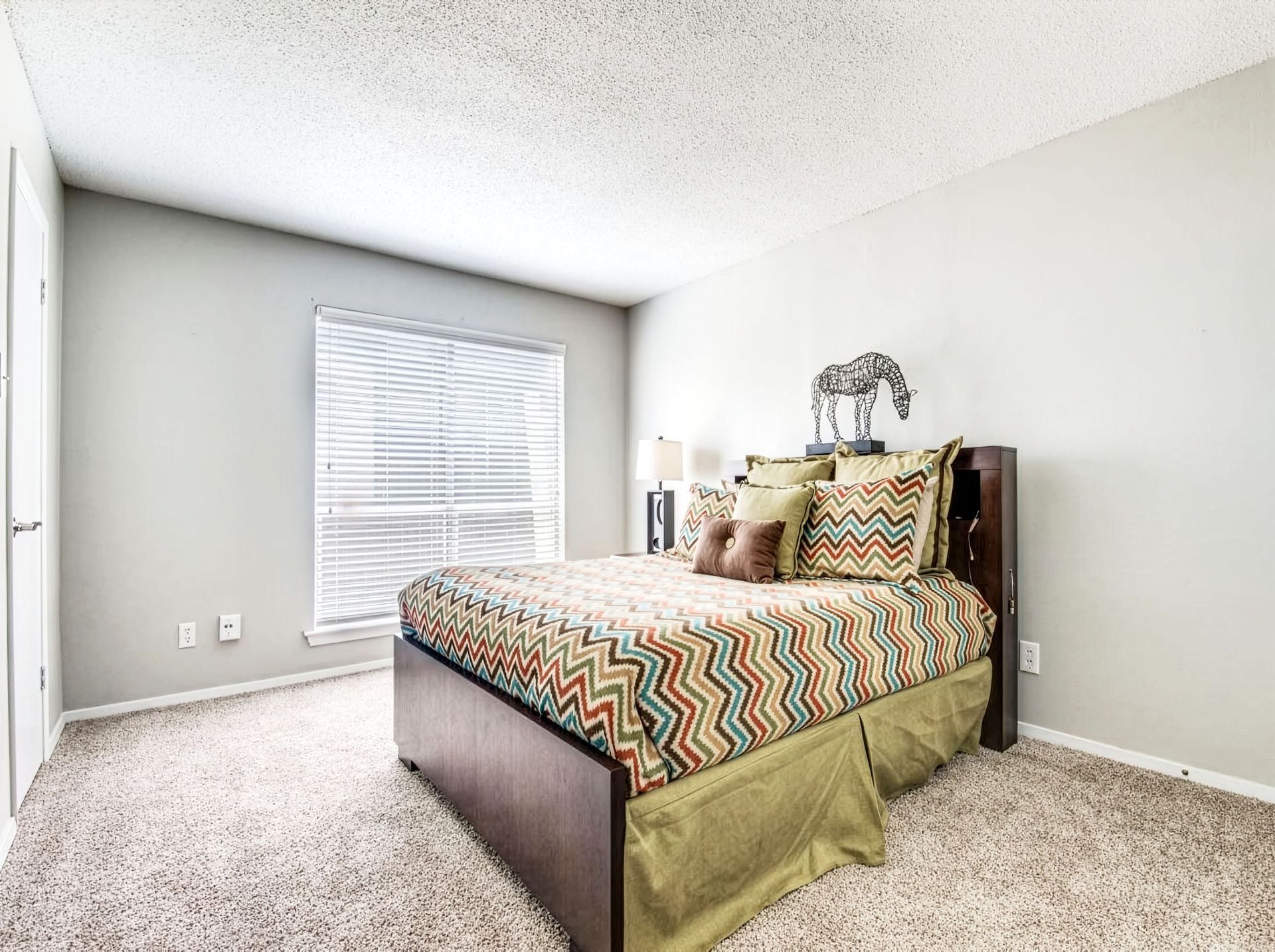 Bright bedroom with a bed, chevron bedding, carpet, and a window with blinds