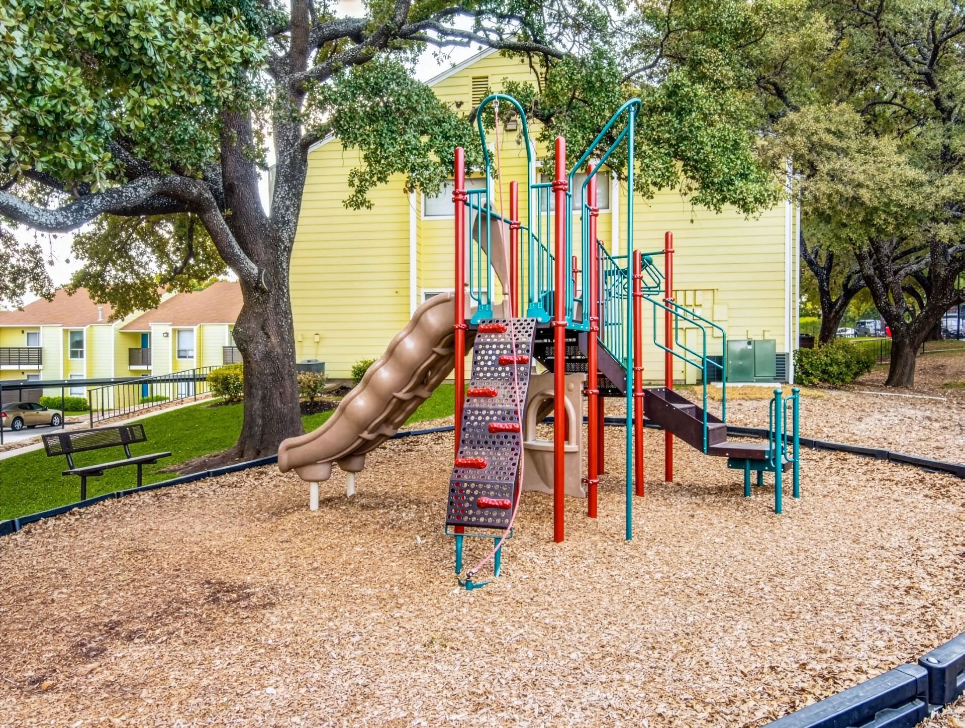 Colorful playground climbing structure and slide in a woodchip courtyard beside yellow apartment buildings.