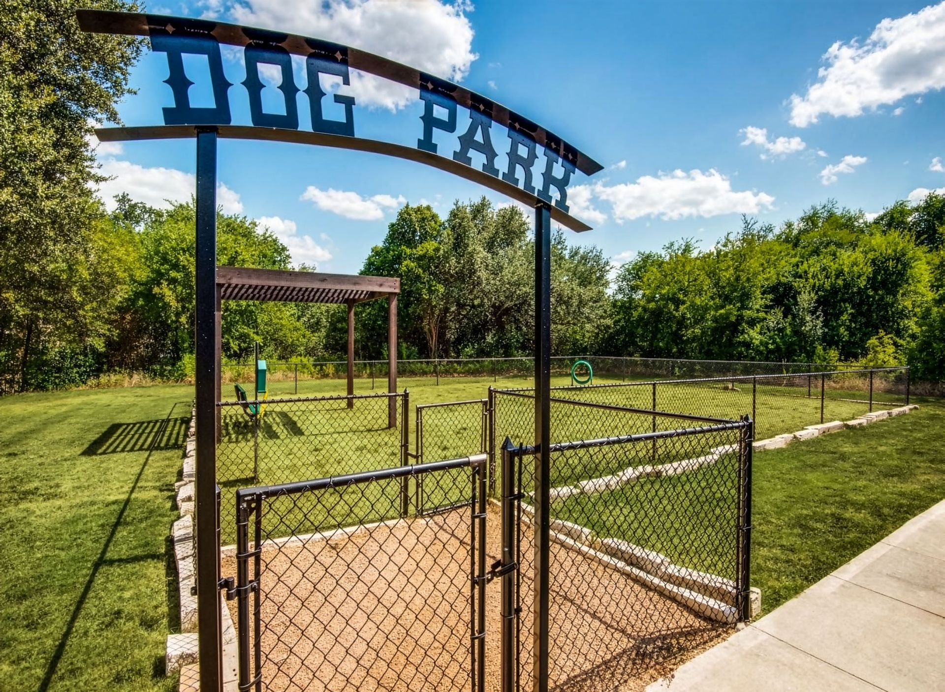 Dog park entrance with black gate, grassy play area, and trees under a blue sky