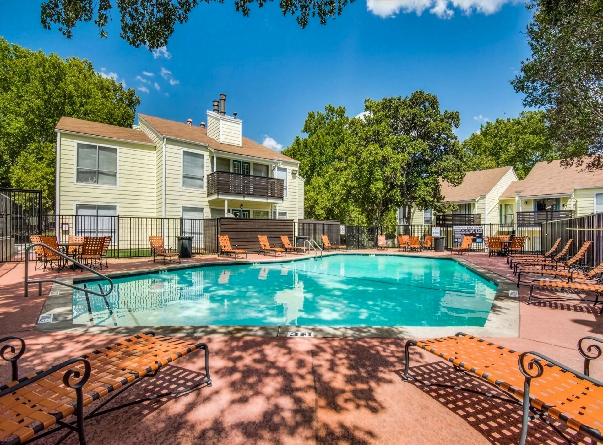 Apartment complex pool courtyard with turquoise water, lounge chairs, and trees under a clear blue sky