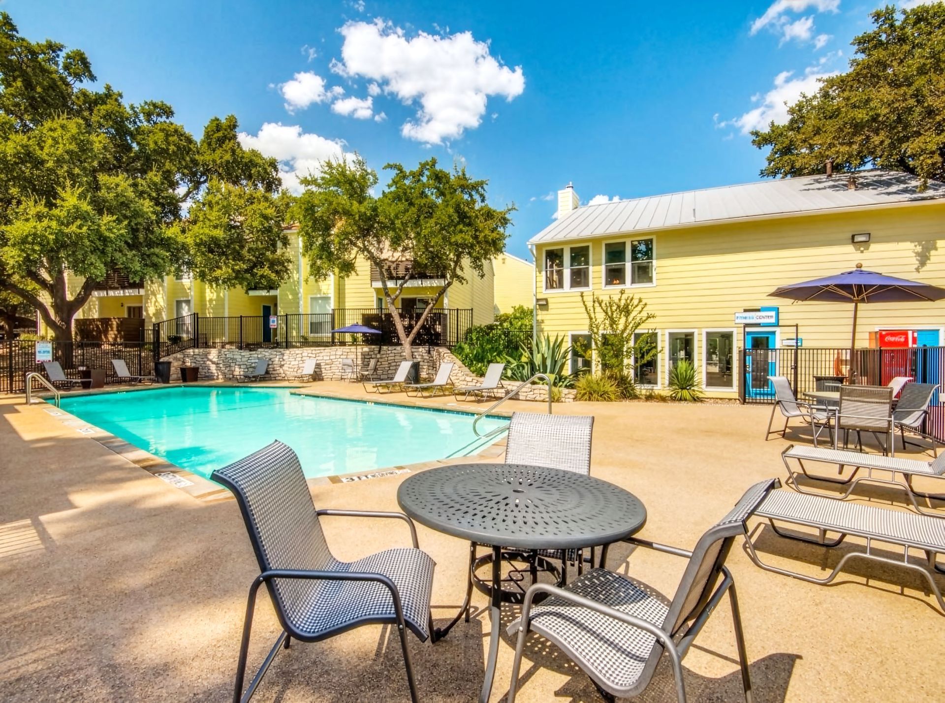 Apartment pool courtyard with turquoise water, lounge chairs, and patio tables under sunny trees