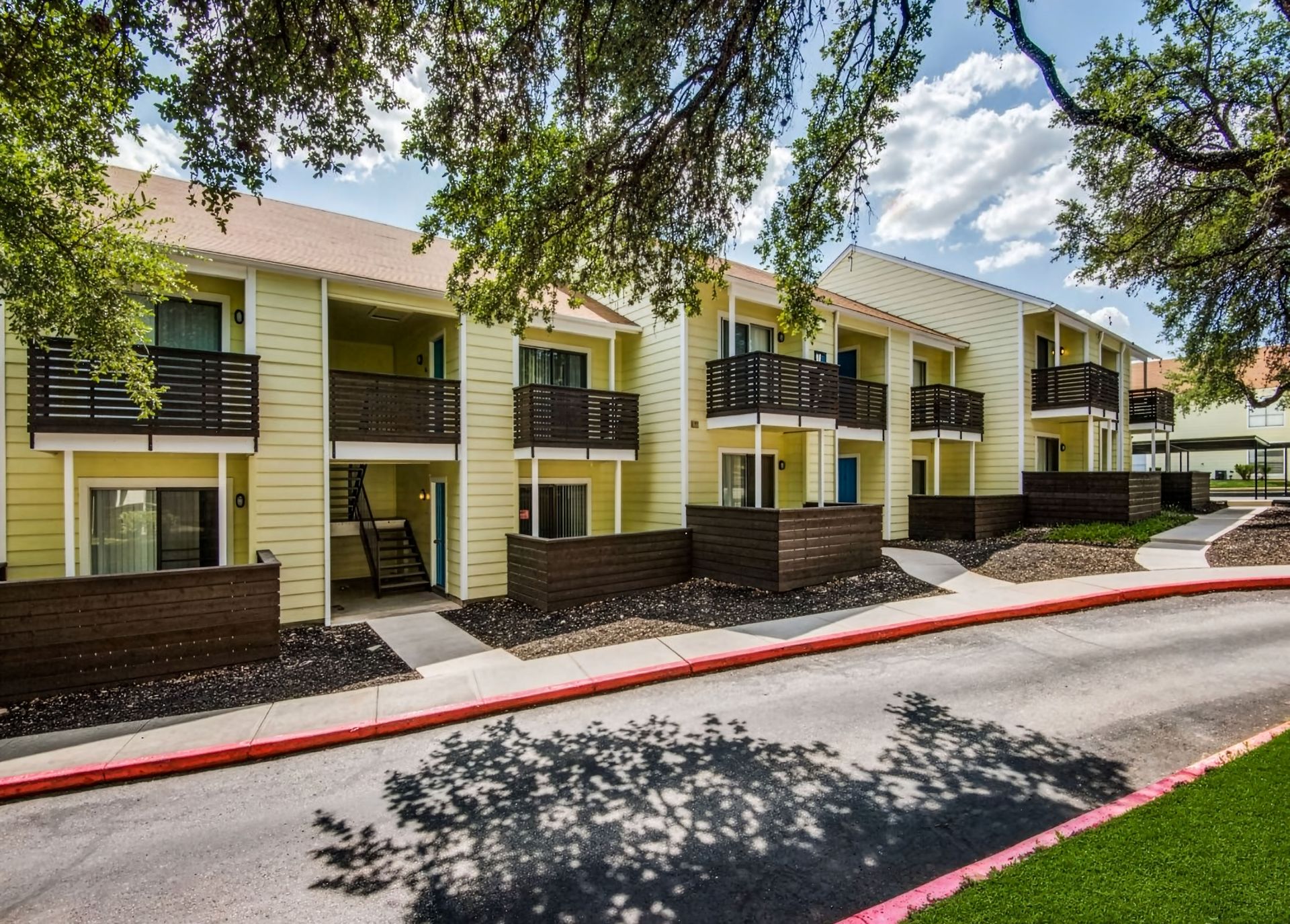 Apartment building with yellow siding, dark railings, and balconies along a tree-lined sidewalk