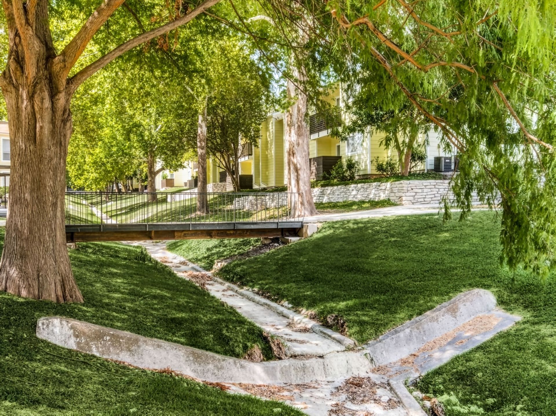 Shaded park with a small stream, footbridge, and green grassy banks under trees