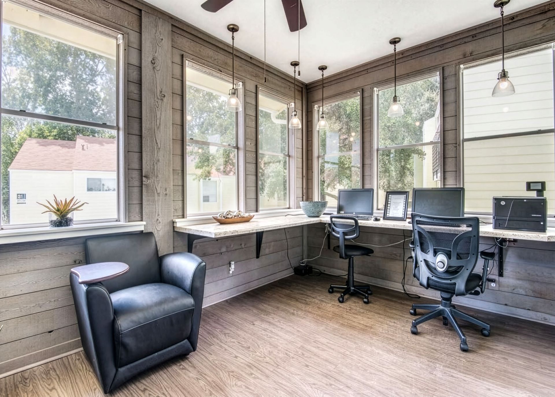 Bright sunroom office with large windows, two desks, rolling chairs, and a black armchair
