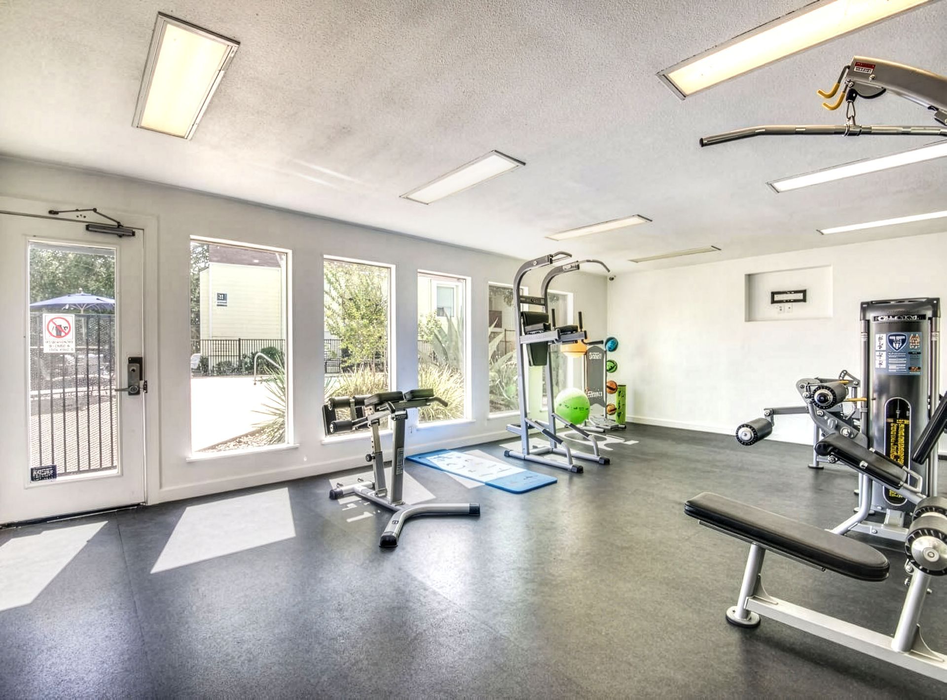 Bright gym room with exercise machines, benches, and large windows letting in natural light