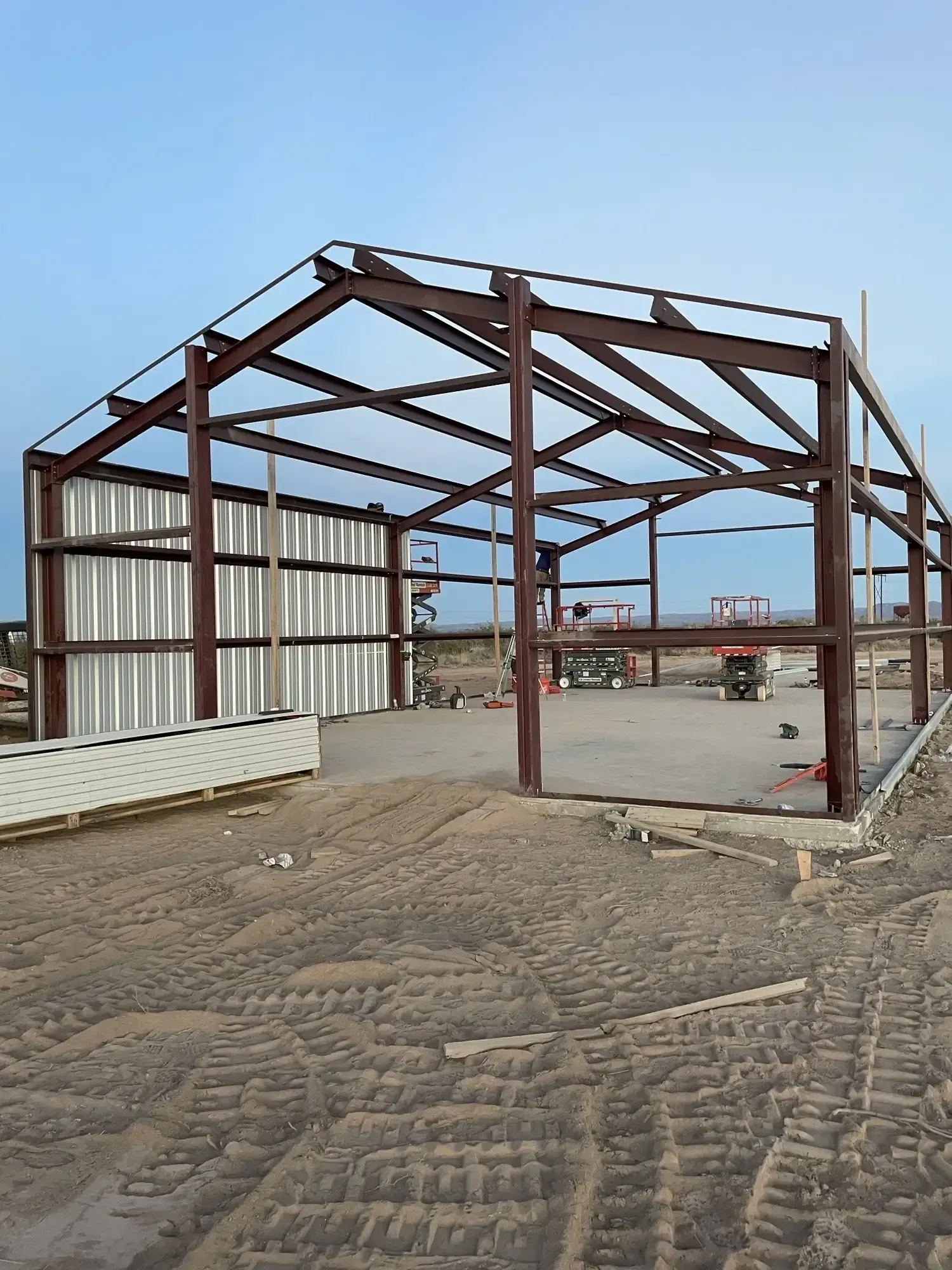 Steel frame of a building under construction, with corrugated metal siding and a concrete foundation in a dirt lot.