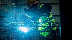 Welder wearing a safety mask and vest working on metal, with sparks flying in a dim industrial setting.