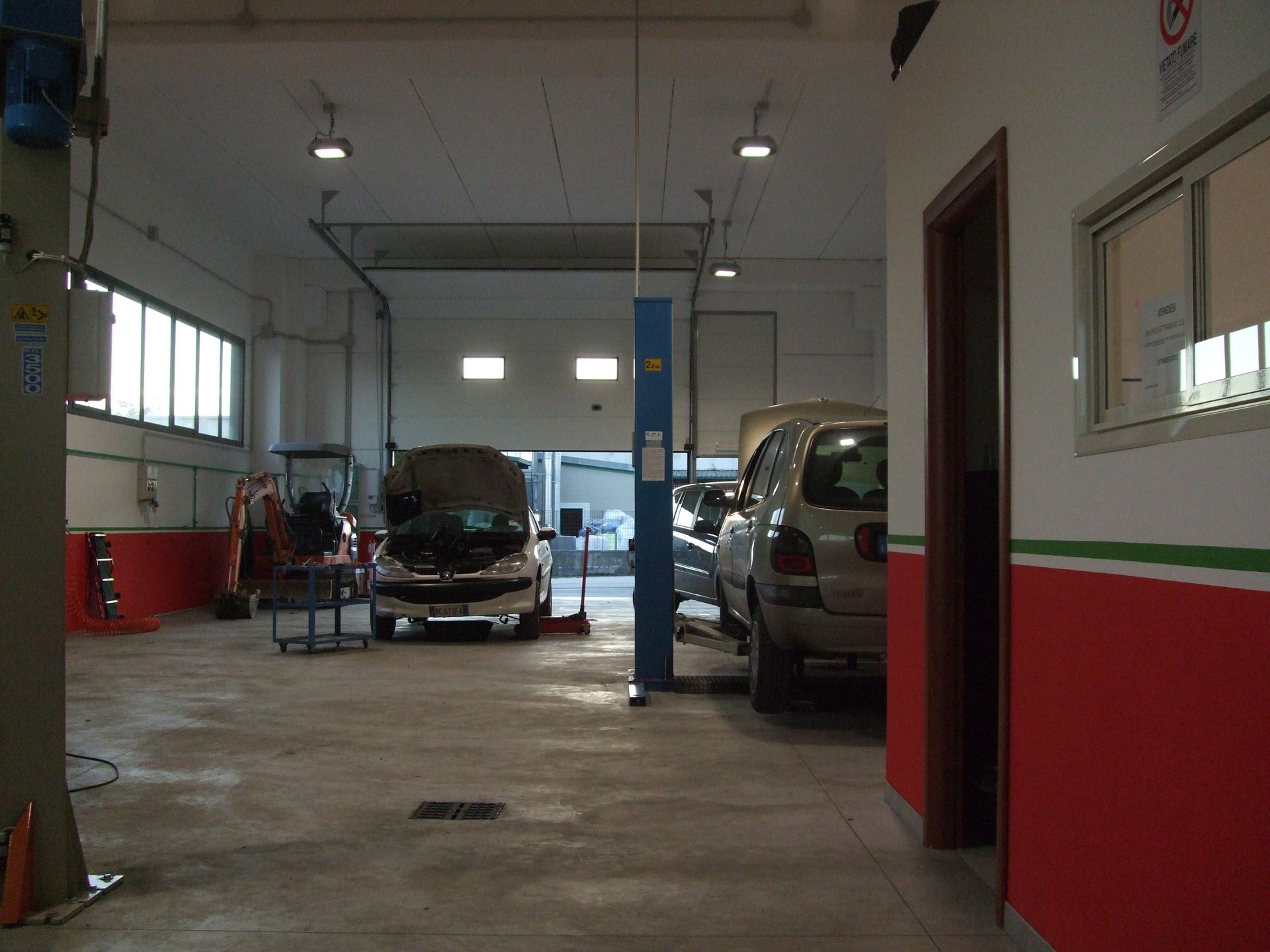 Interno di un'officina con auto su ponti sollevatori, attrezzi e portelli di carico aperti.