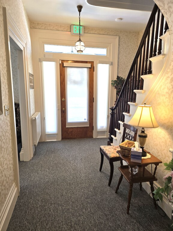Narrow hallway with carpet, staircase, wooden door, and a small table with lamp and chairs