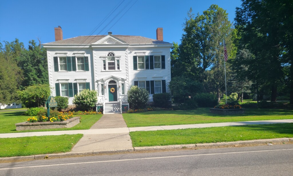 White two-story colonial house with a front porch, lawn, and flower beds under a blue sky