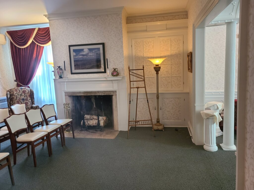 Elegant parlor with fireplace, chairs, draped window, and a lamp beside a doorway.