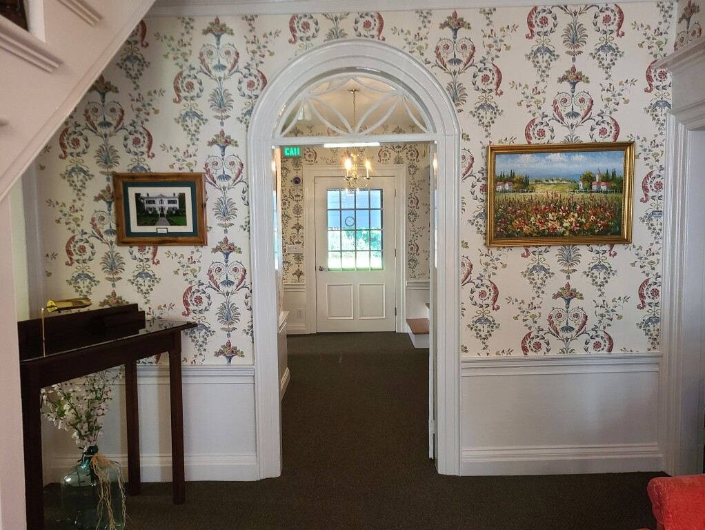Hallway with floral wallpaper, framed paintings, and an arched doorway leading to a bright door at the far end