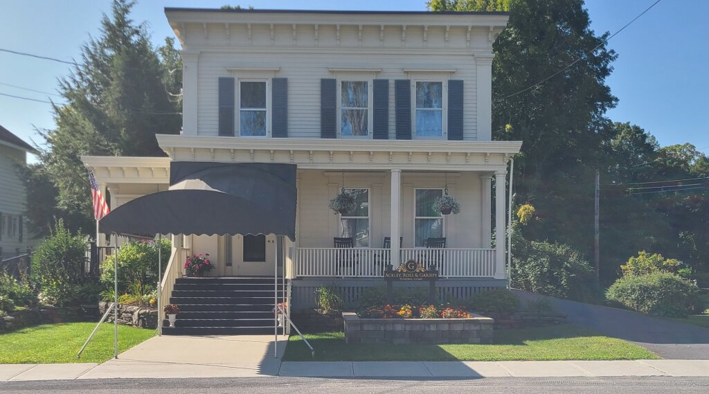 Two-story white house with blue shutters, front porch, black awning, and manicured lawn in daylight