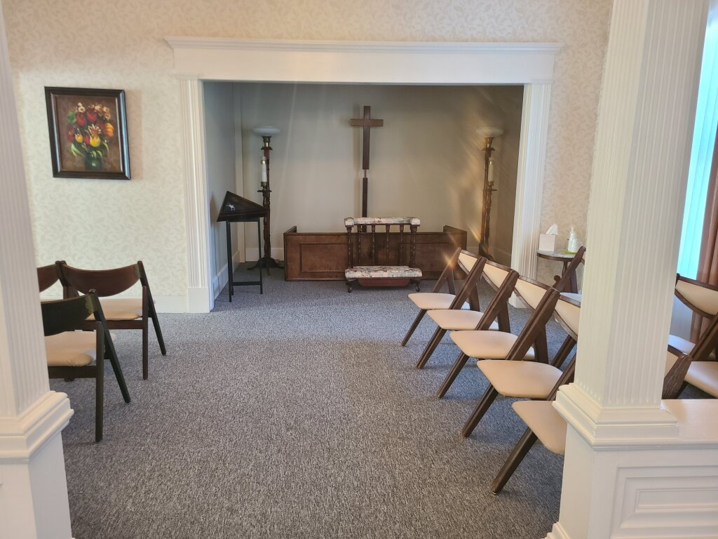 Small chapel room with altar, cross, and rows of folding chairs on carpet.