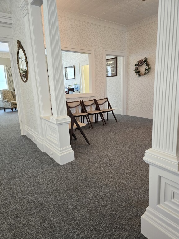 Empty carpeted hallway with white columns, chairs, and a wreath on the wall