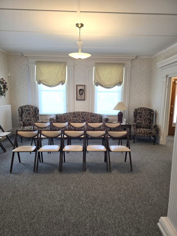 Formal living room with floral sofas, matching armchairs, and a row of chairs facing the camera beneath two windows.