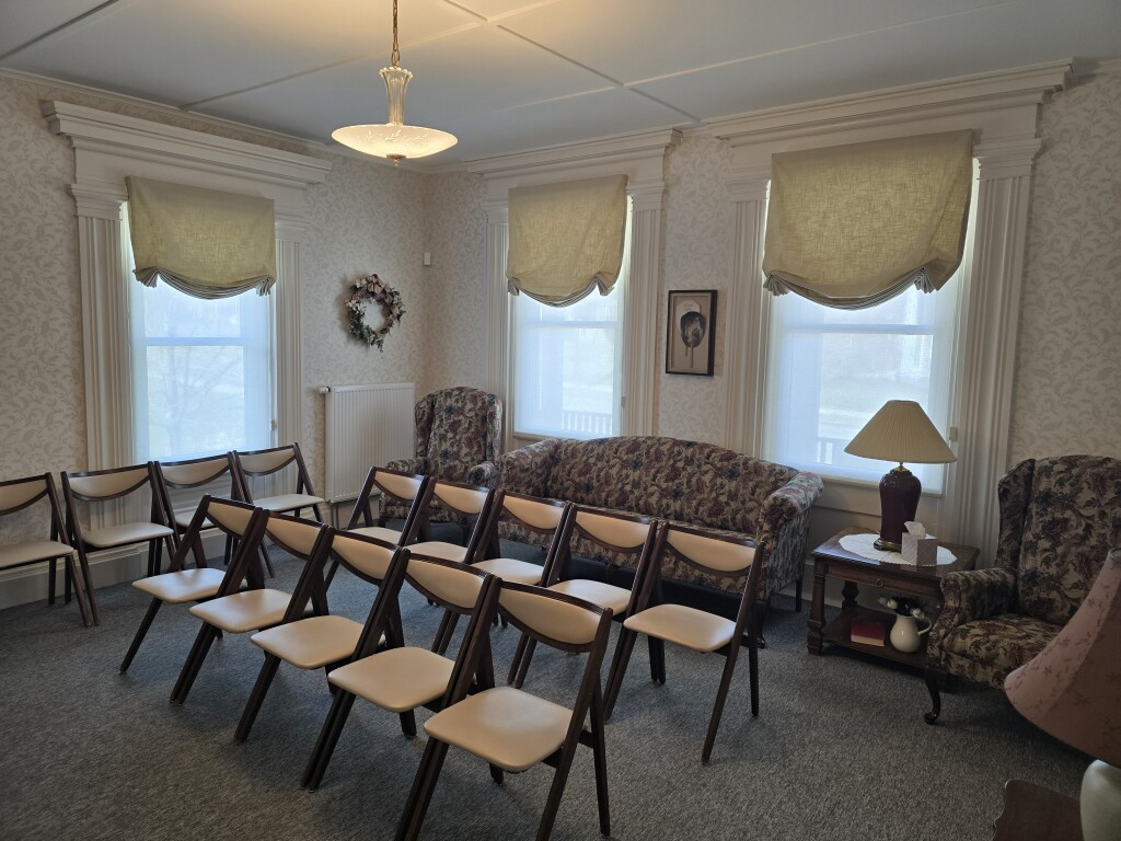 Empty meeting room with rows of folding chairs facing the front, patterned sofas, and three windows with shades.