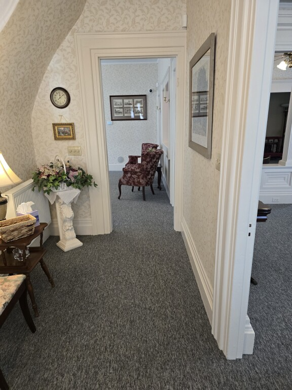 Narrow carpeted hallway with white walls, framed art, and chairs in a home interior.