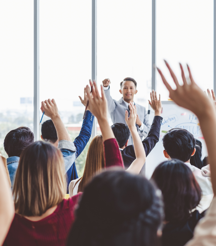 Audience raising hands toward a speaker in a bright conference room with large windows