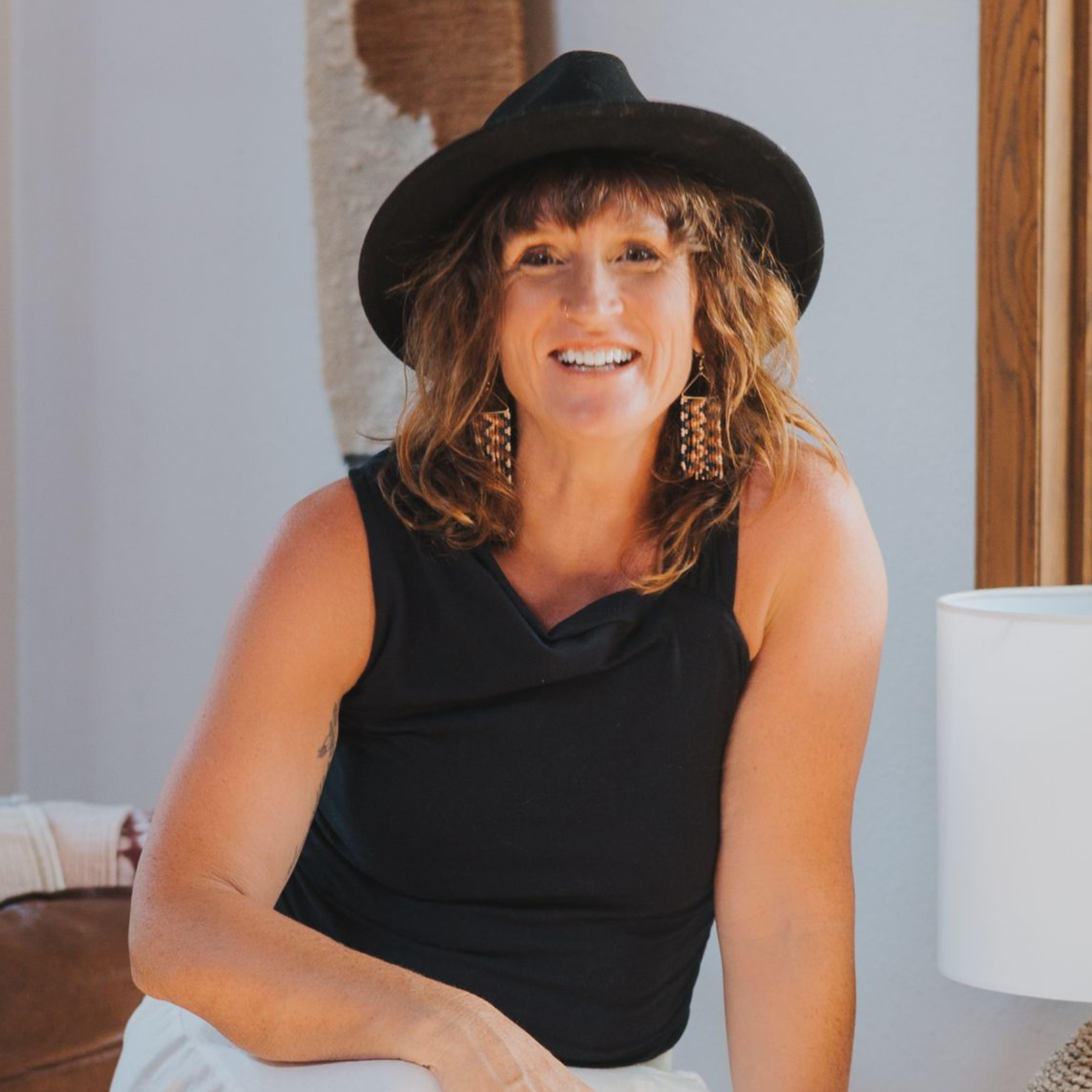 Woman in black hat and sleeveless top sitting indoors, smiling beside a white lamp