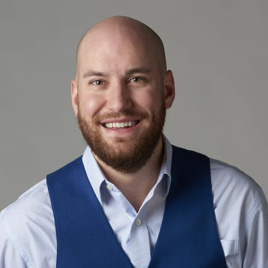 Smiling bald man with a beard in a blue vest and light shirt against a gray background