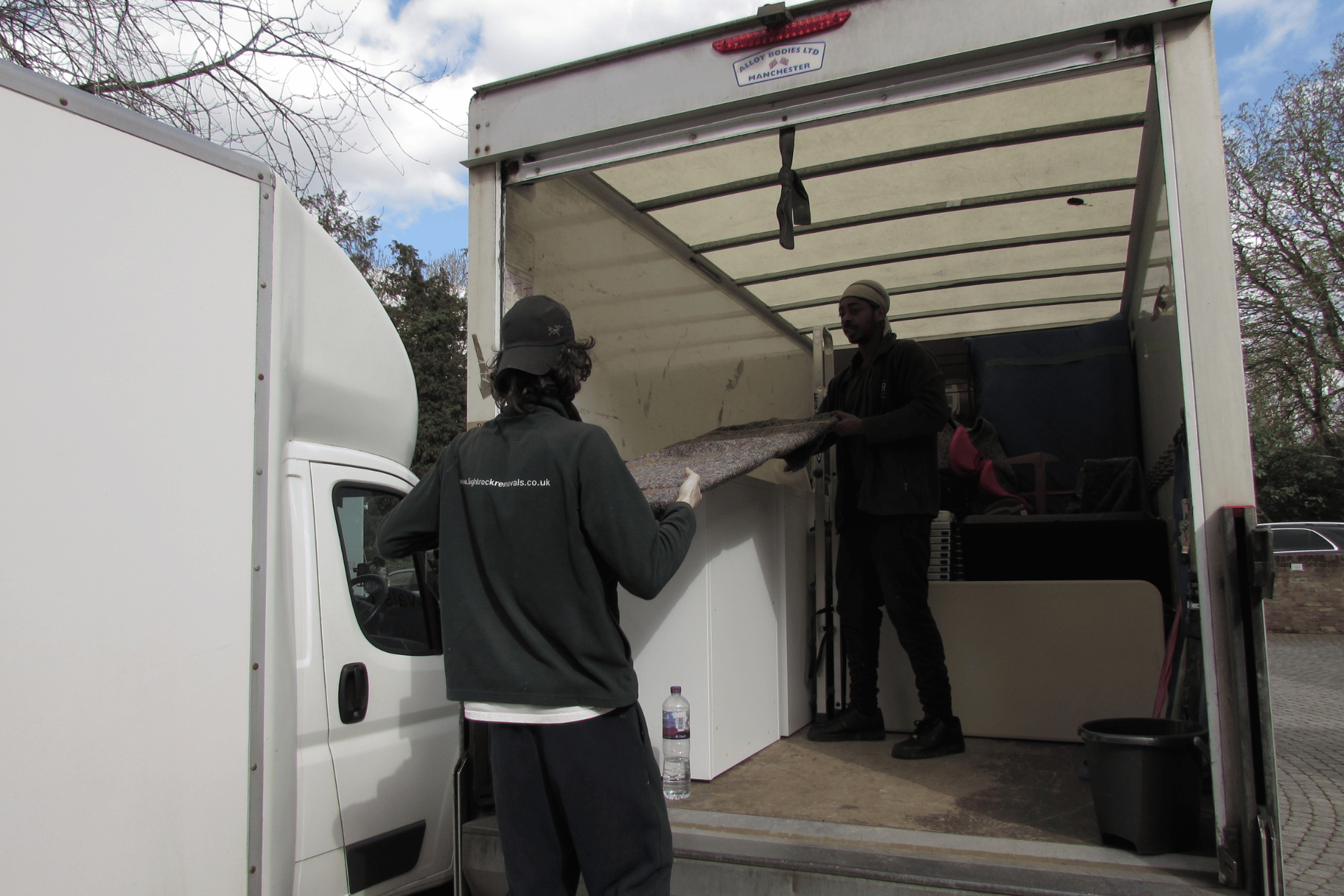 Two people loading a white refrigerator into a large moving truck outside.
