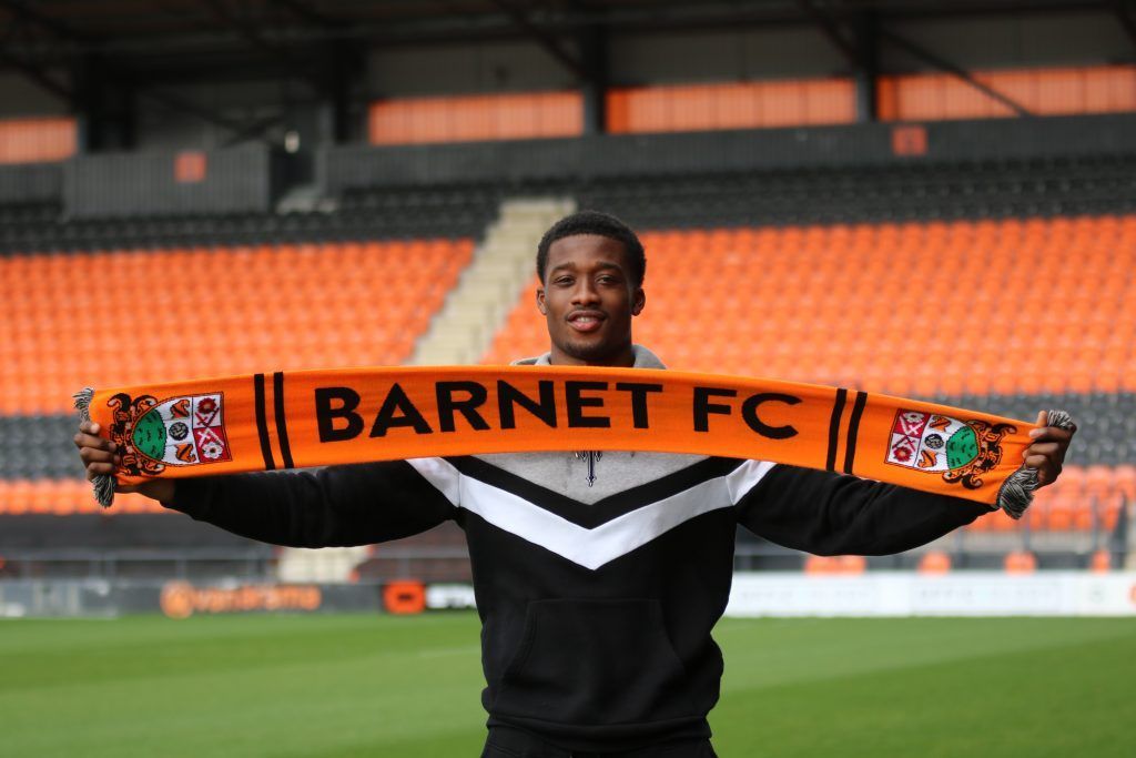 Footballer holding a Barnet FC scarf, smiling in front of an orange and black stadium.