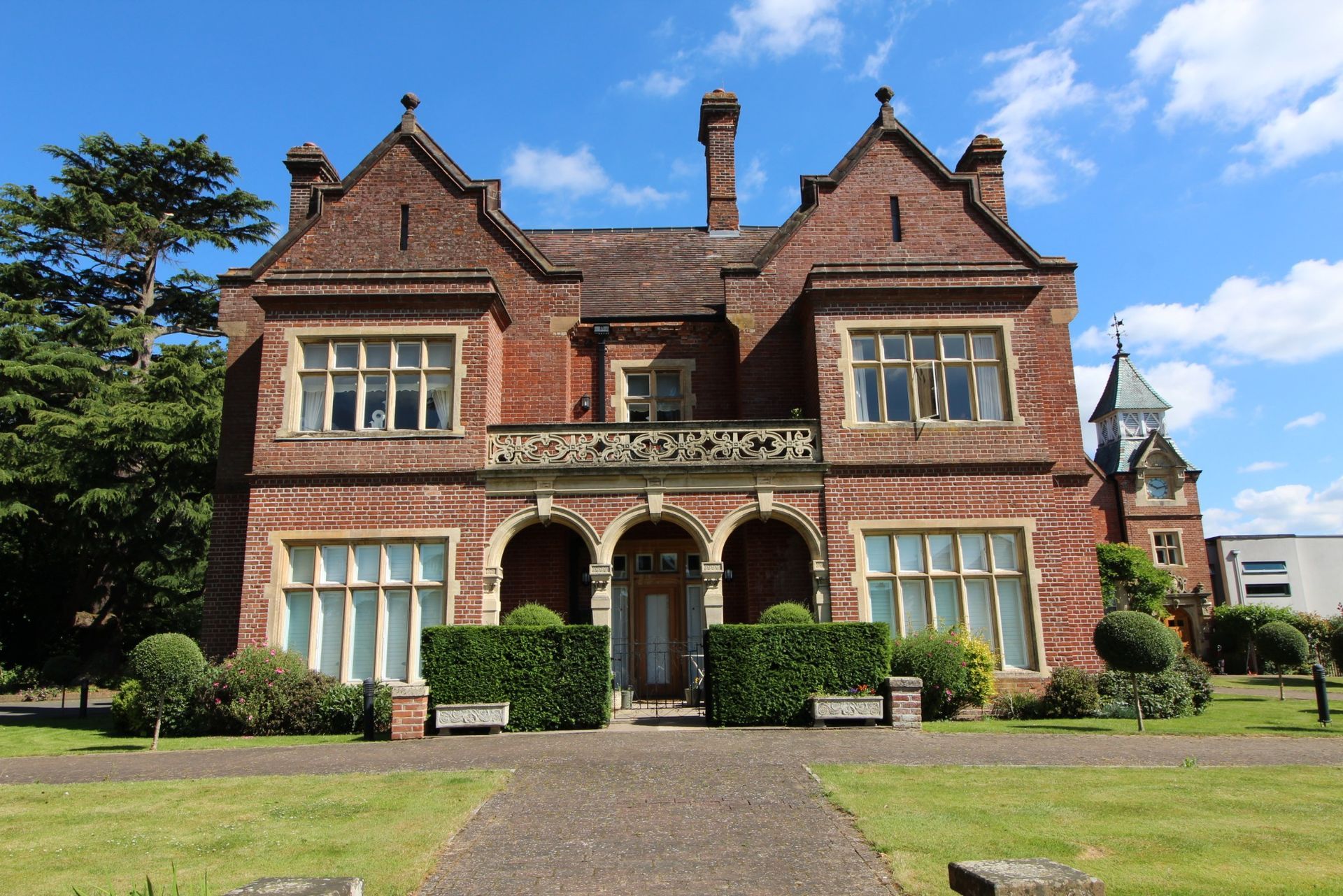 Red brick Victorian manor house with gables, a balcony, and a small tower, set in a grassy landscape.