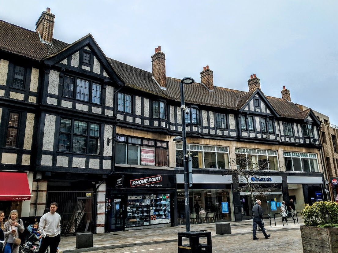 Tudor-style buildings with shops on a cobblestone street, people walking.