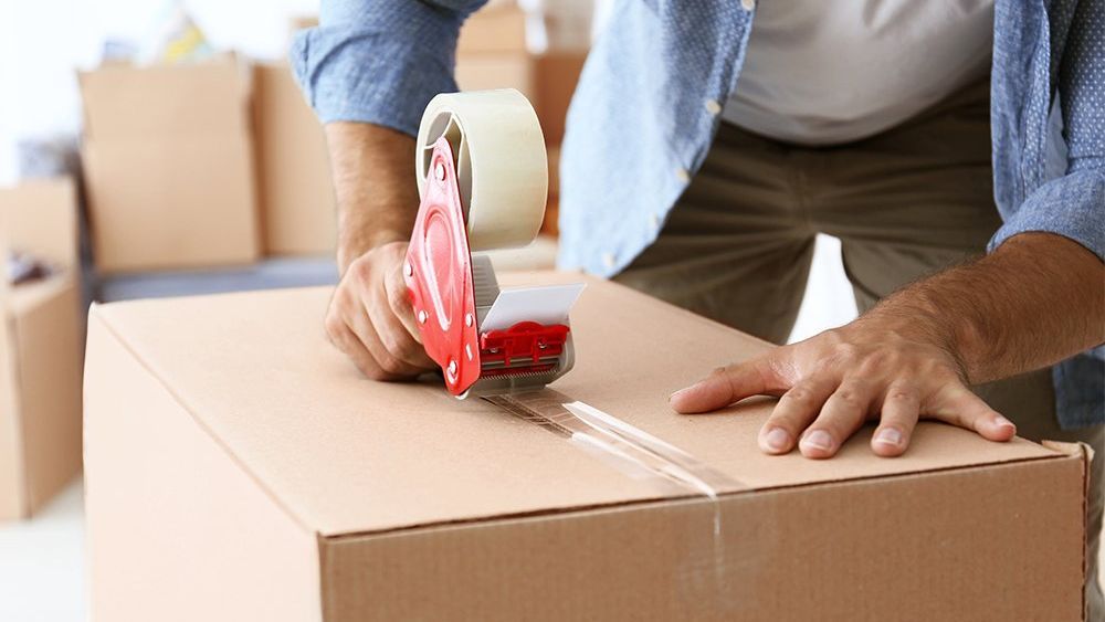 Person sealing a cardboard box with packing tape, preparing for a move.