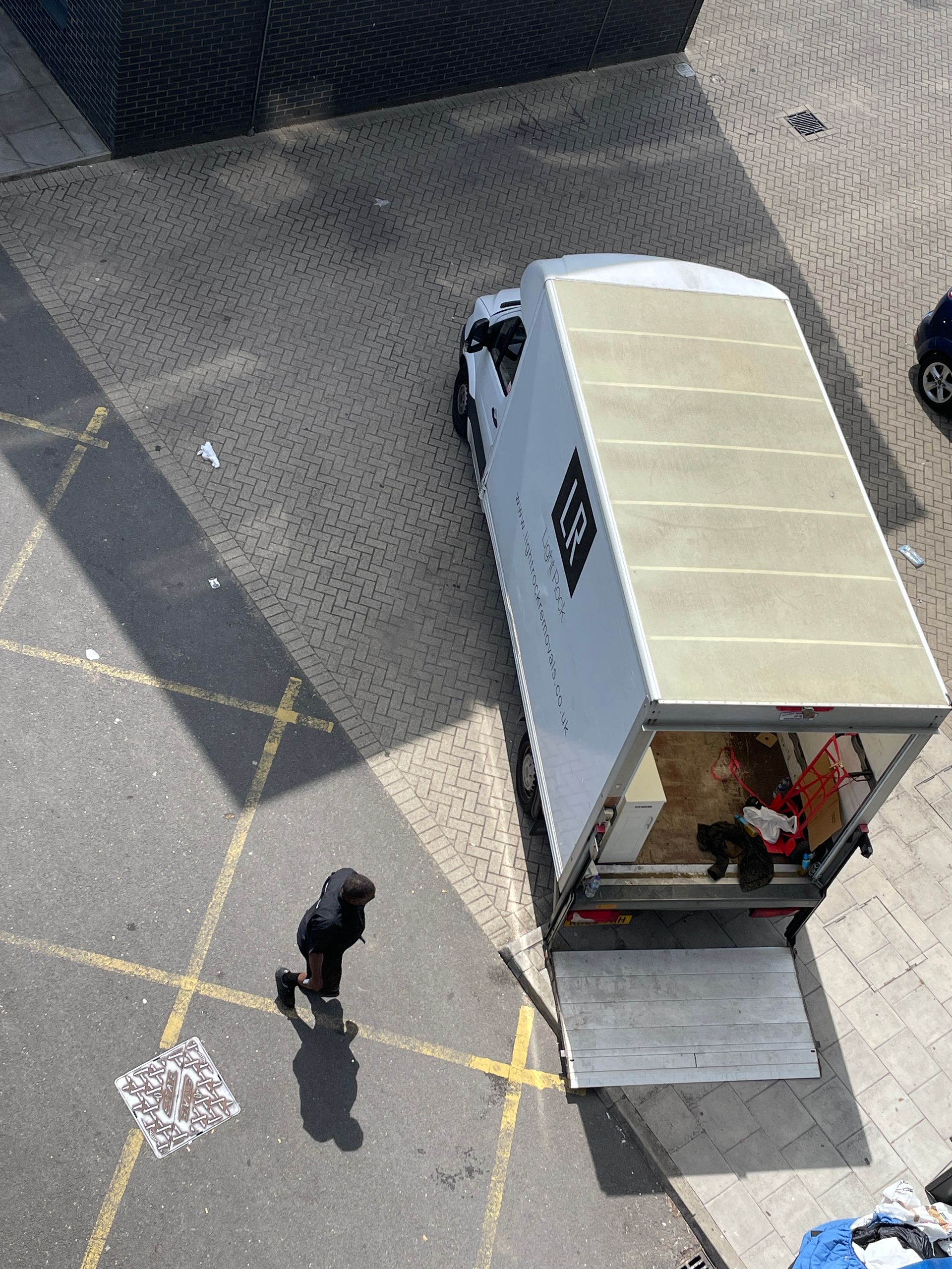 Overhead view of a white delivery truck parked on pavement with a lowered ramp. A person in dark clothing walks past.