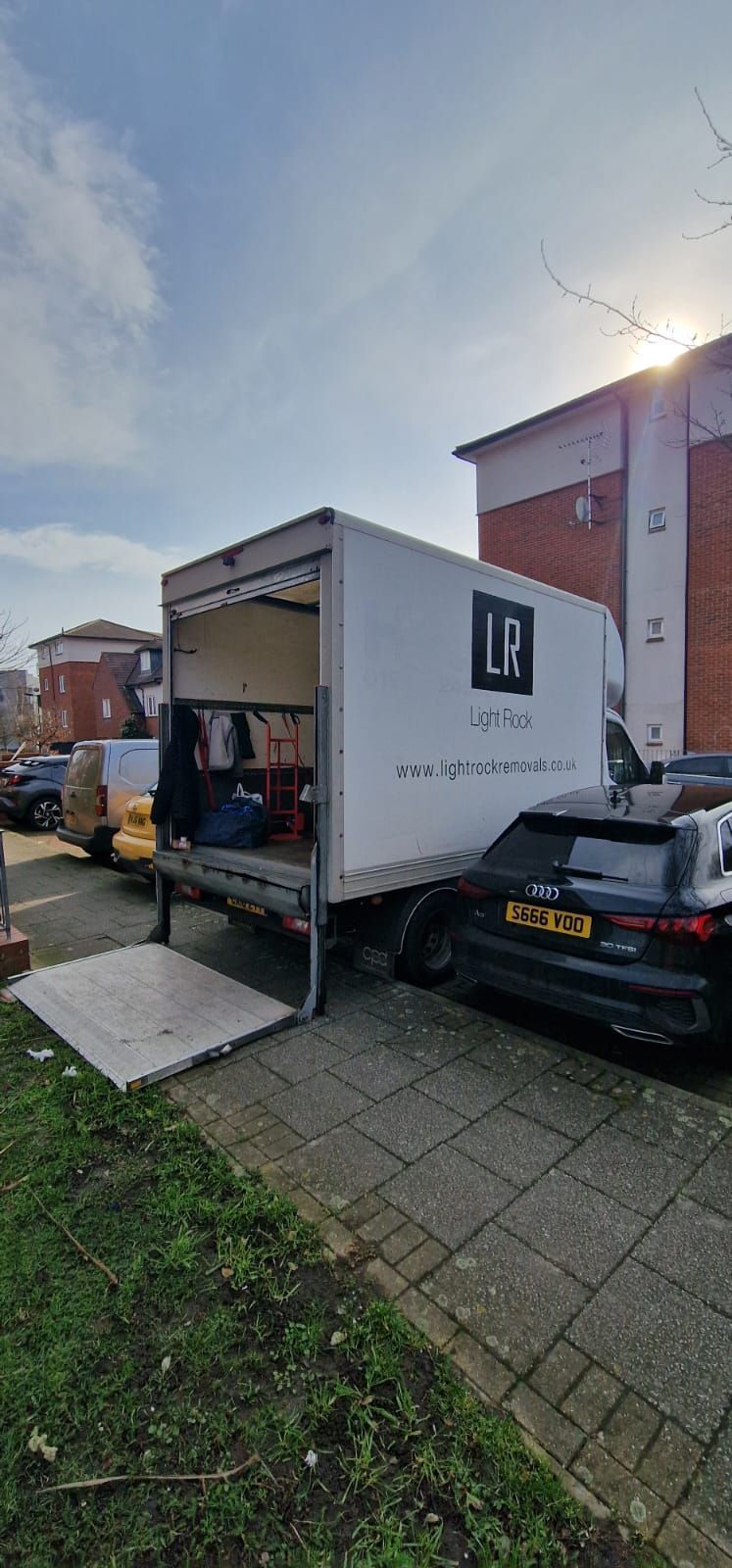 A moving truck with an open ramp parked on a paved area next to a building. The truck has the letters