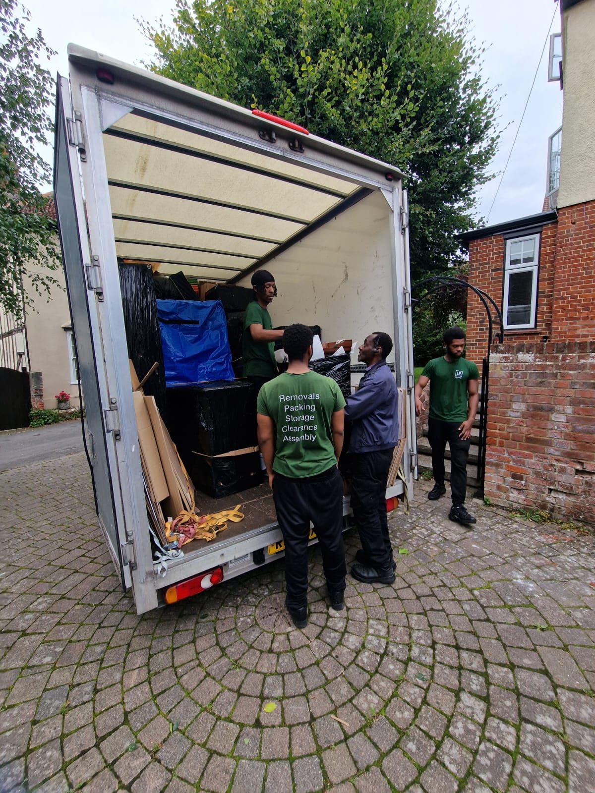 They stand on a cobblestone drive, wearing green shirts, loading boxes and furniture from a house with a brick wall.