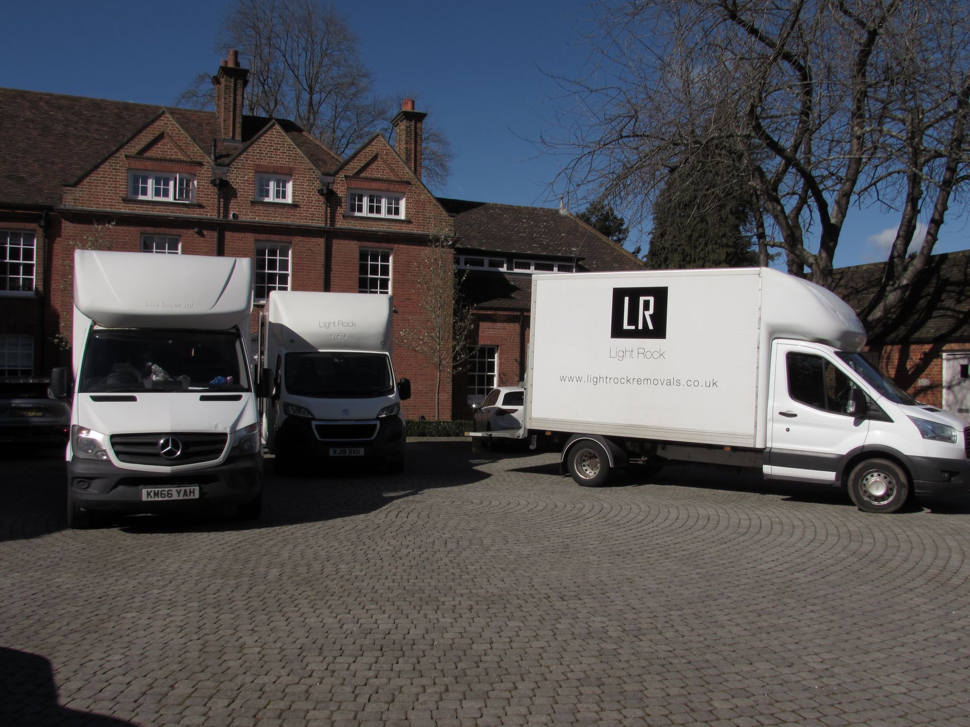 Three white moving vans parked in front of a brick building with a cobblestone parking area under a blue sky.