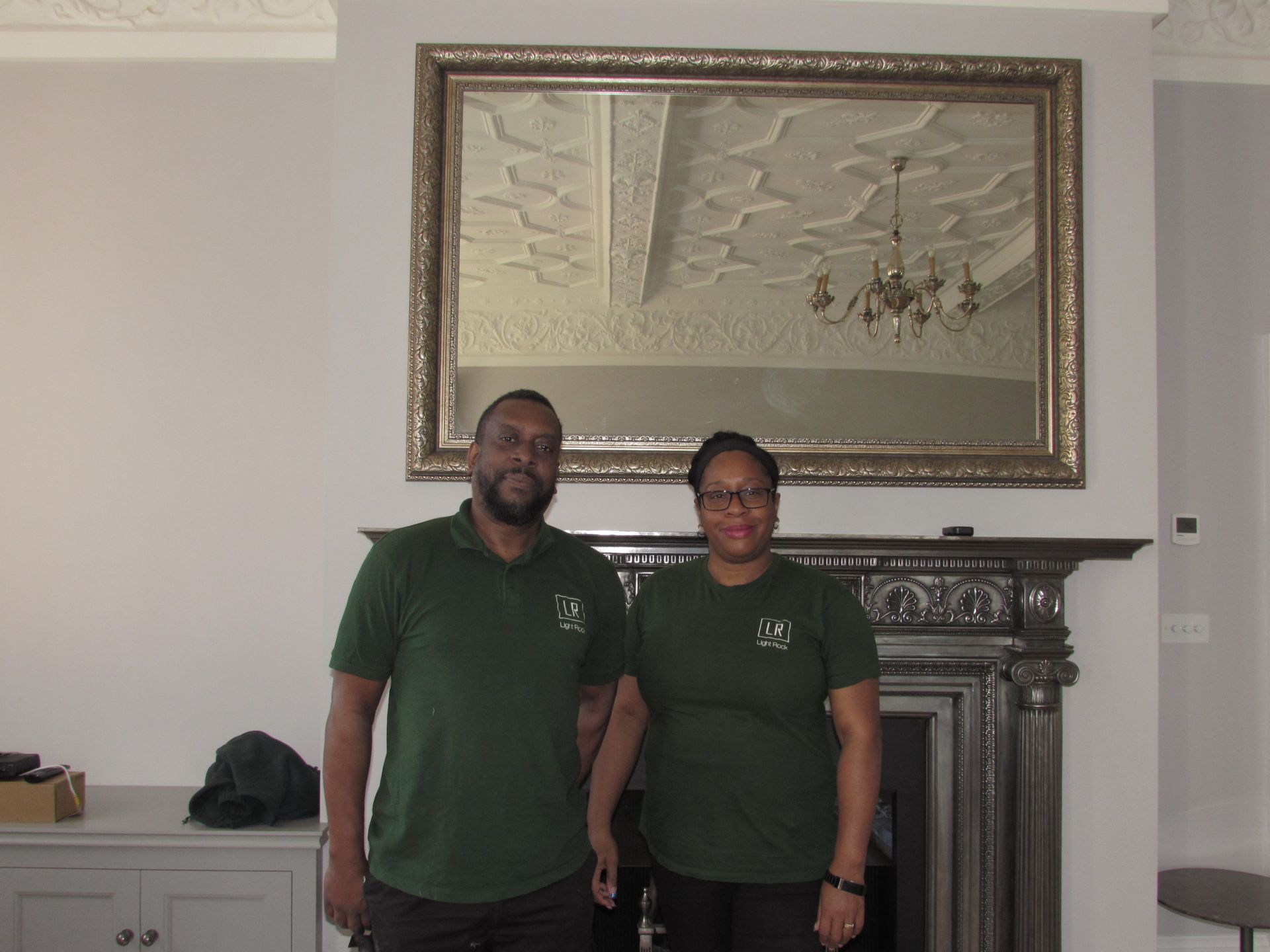 Two people in green shirts stand in front of a fireplace and a large mirror. The mirror reflects a chandelier