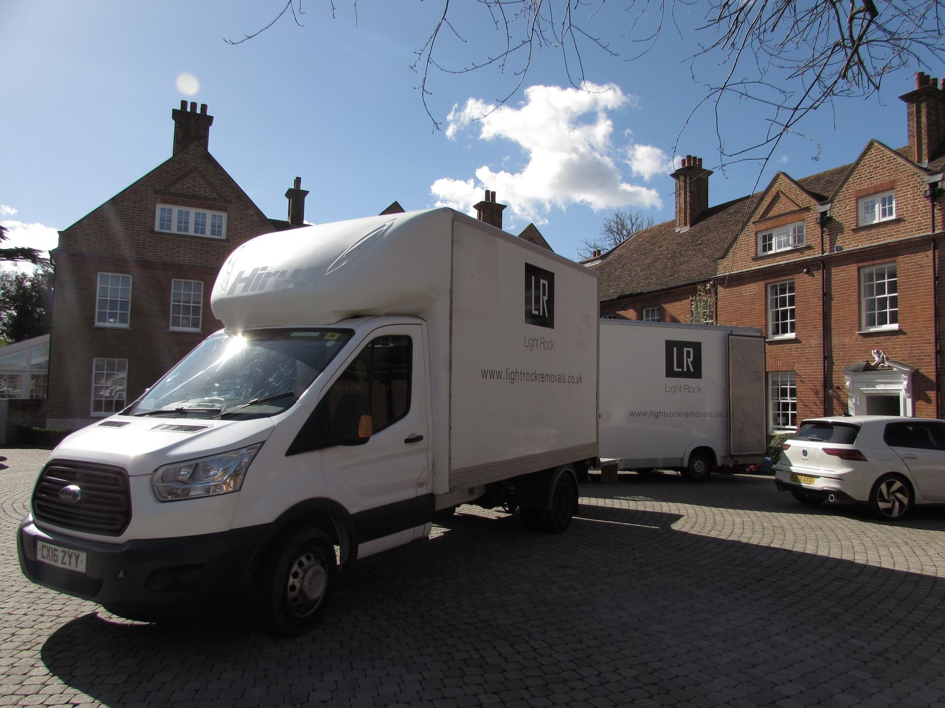 White moving vans parked on a cobblestone driveway in front of a large brick building on a sunny day.