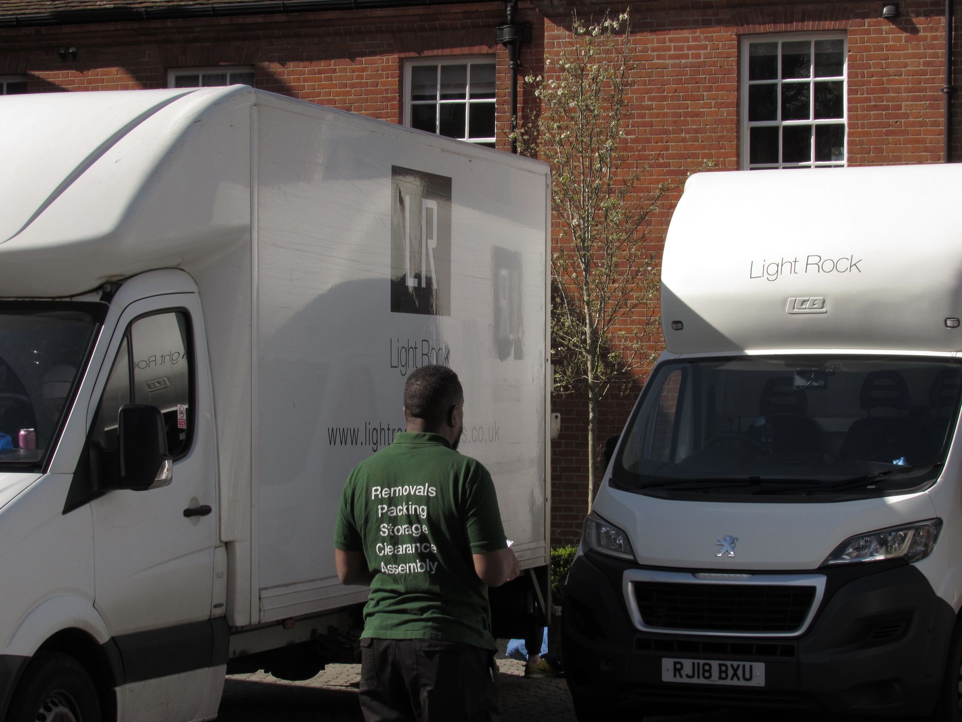 Man in green shirt stands between two white moving vans, in front of a brick building.