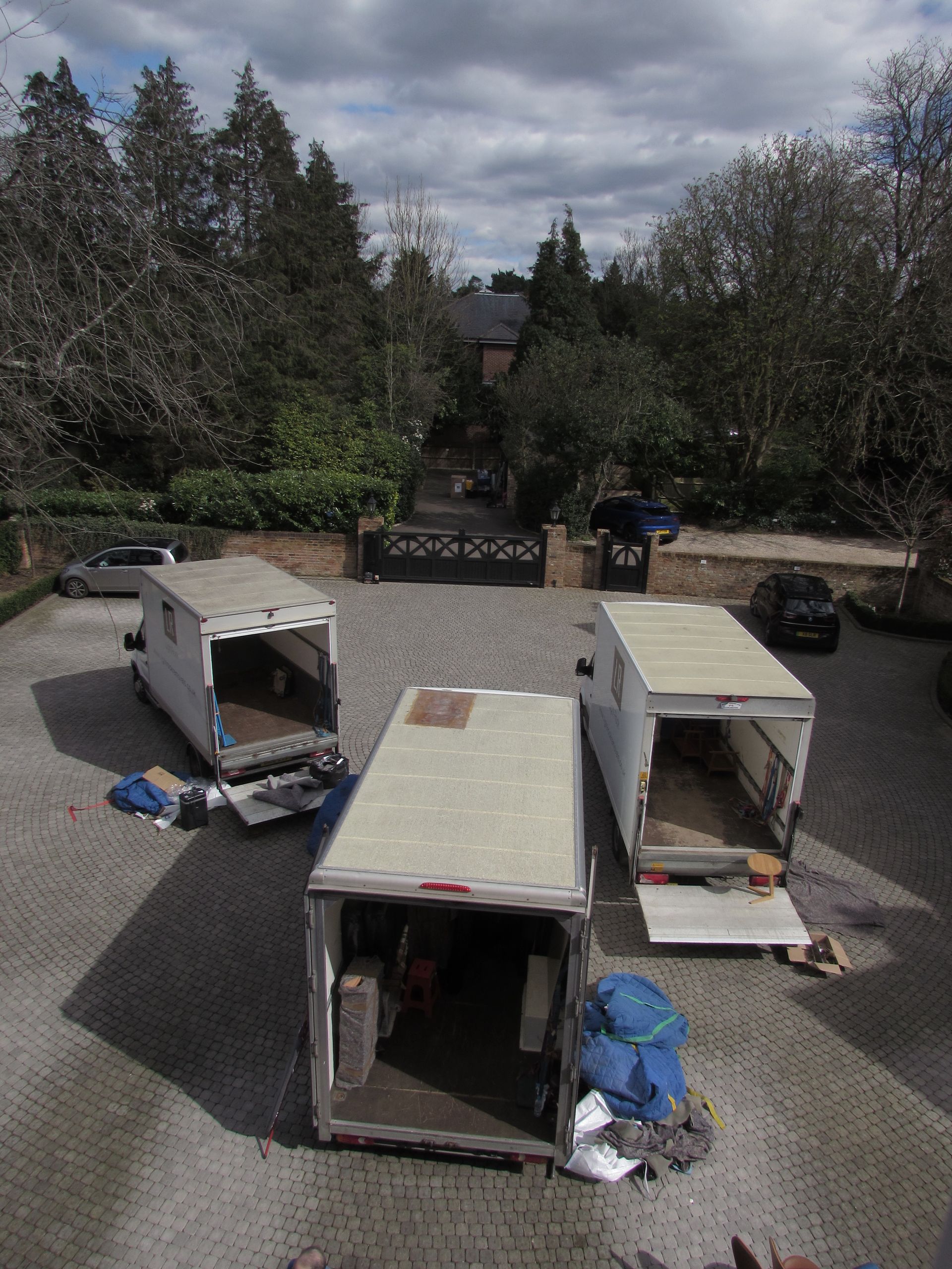 Three moving trucks with open backs in a driveway of a large house. Some belongings are on the ground.