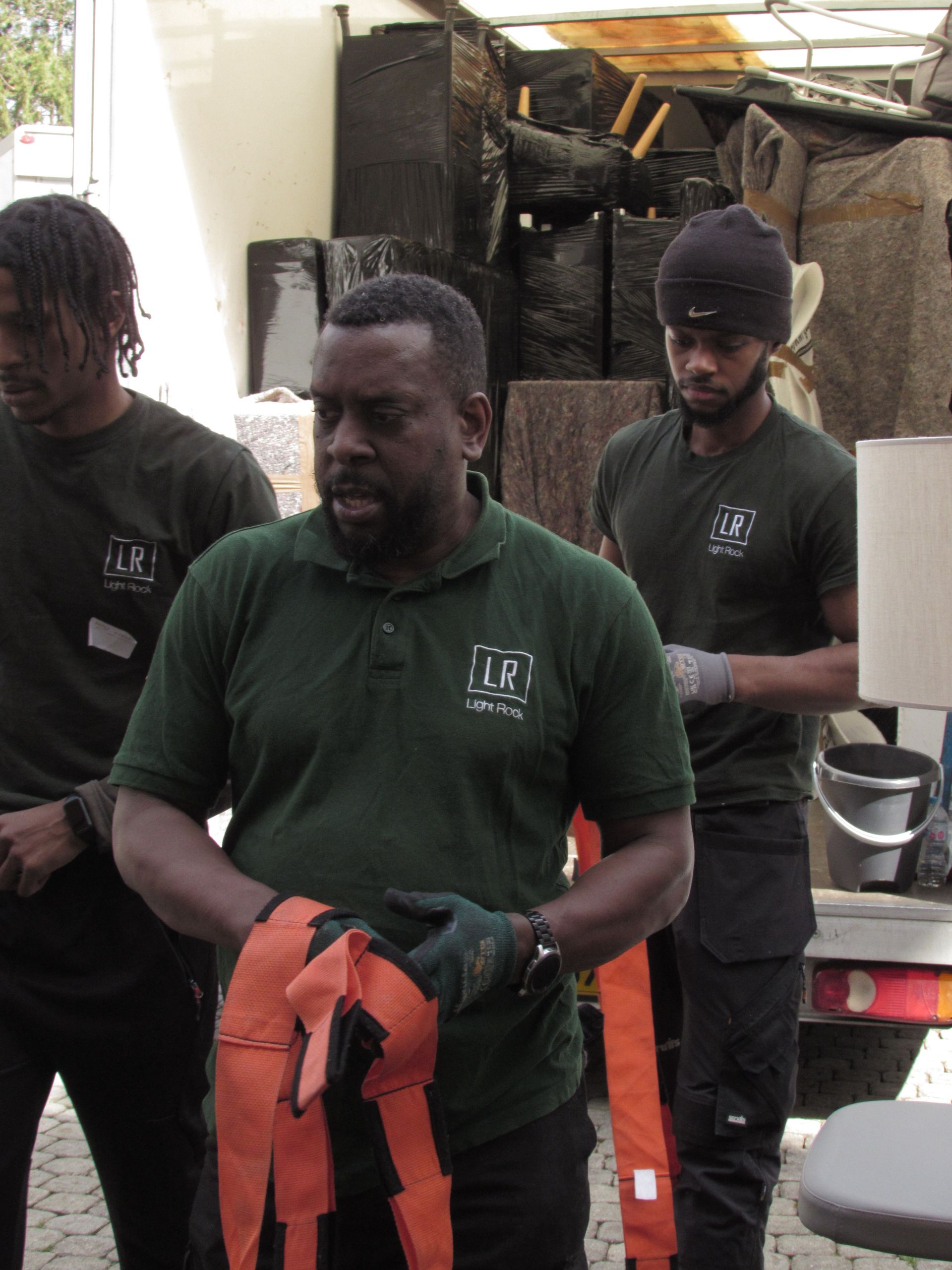 Three movers in green shirts loading a truck. The central man, holding orange straps, speaks with a serious expression.