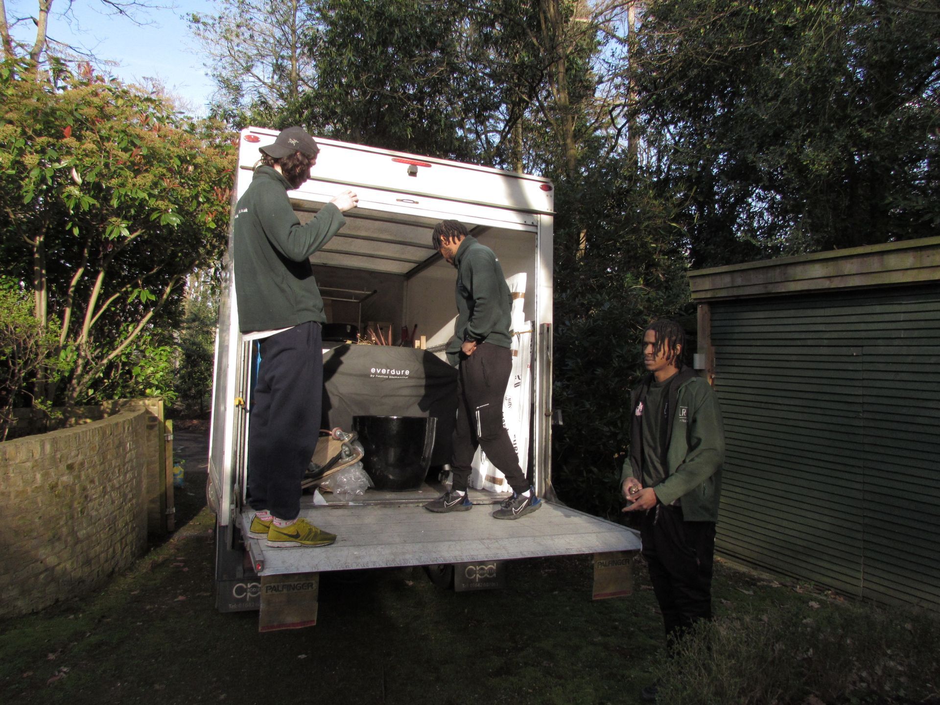 Three people loading items into a white box truck outdoors. One person stands inside the truck, another in the doorway,