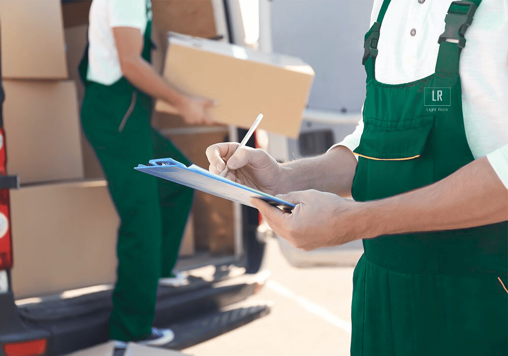 Two movers load boxes into a van; one in blue overalls writes on a clipboard, the other carries a box.