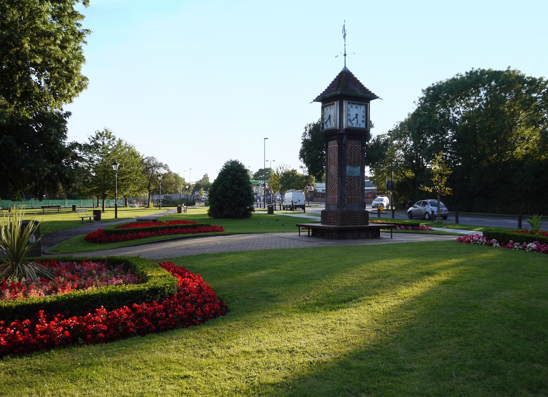 A clock tower in a park with red flowers and a grassy lawn under a clear sky.