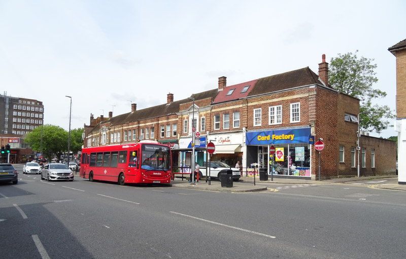 Red bus on a road in front of brick buildings with shops. Cars and a tall building are also present.