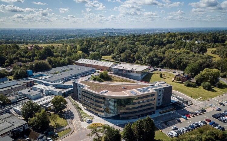 Aerial view of a modern building complex with a curved roof, green space, and surrounding trees.