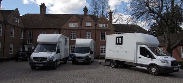 Three white moving vans parked in front of a red brick building with multiple windows and chimneys. Cloudy sky.