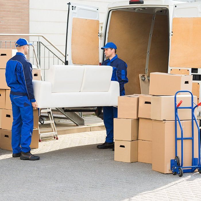 Two movers wearing blue uniforms unload a white couch from a white van. Cardboard boxes are stacked nearby.