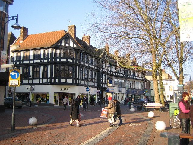 Street scene with Tudor-style building, shoppers crossing brick road, blue sky, trees.