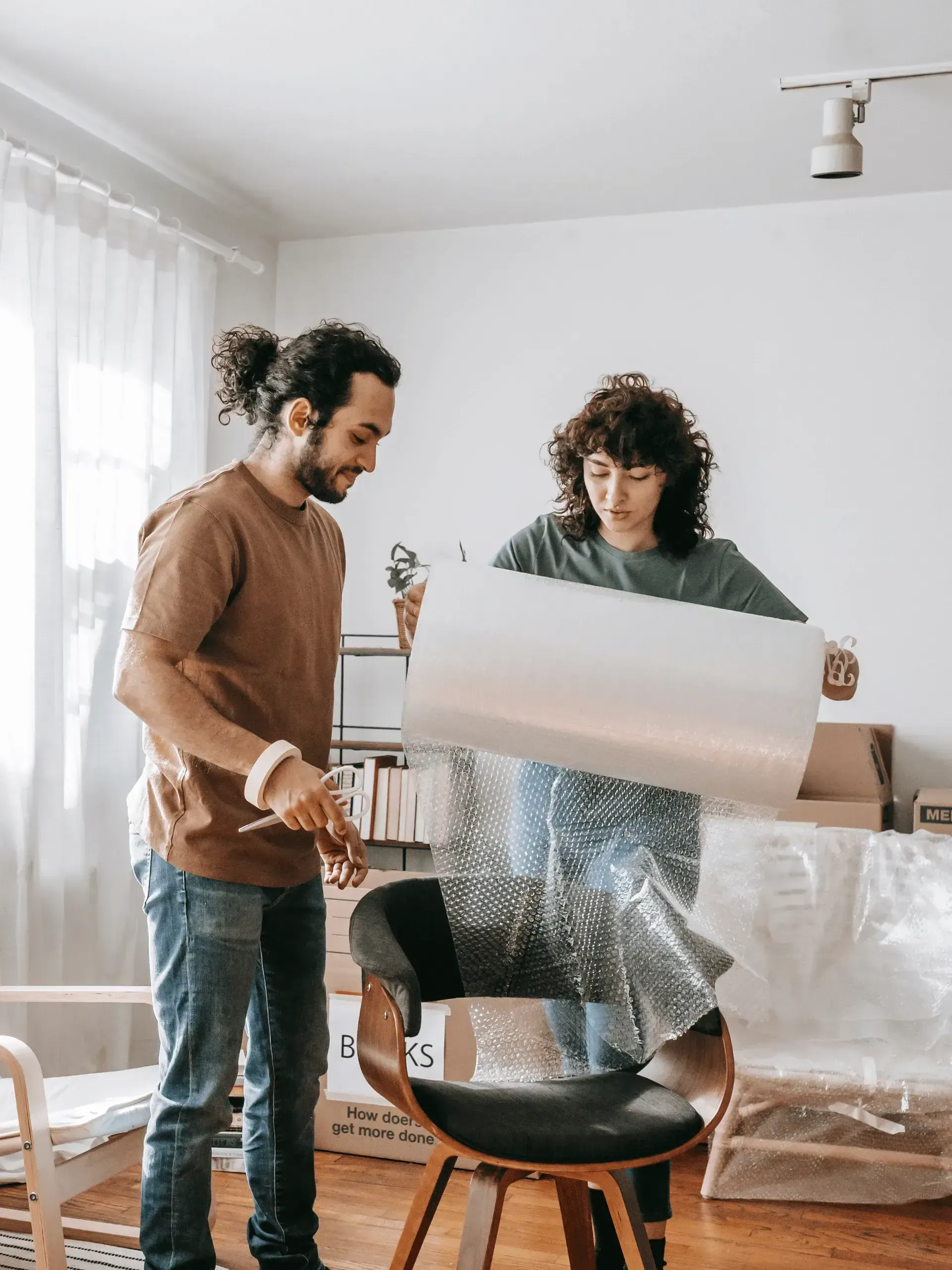 Couple wrapping a chair in bubble wrap, preparing to move; one holds wrap, other holds a knife in a bright, sunny room.