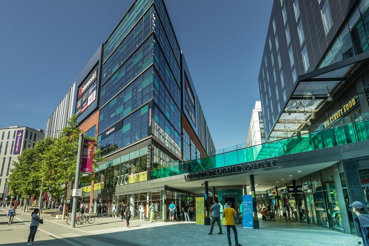 Exterior view of shopping complex, featuring multiple shops building under a bright blue sky. People are walking around.