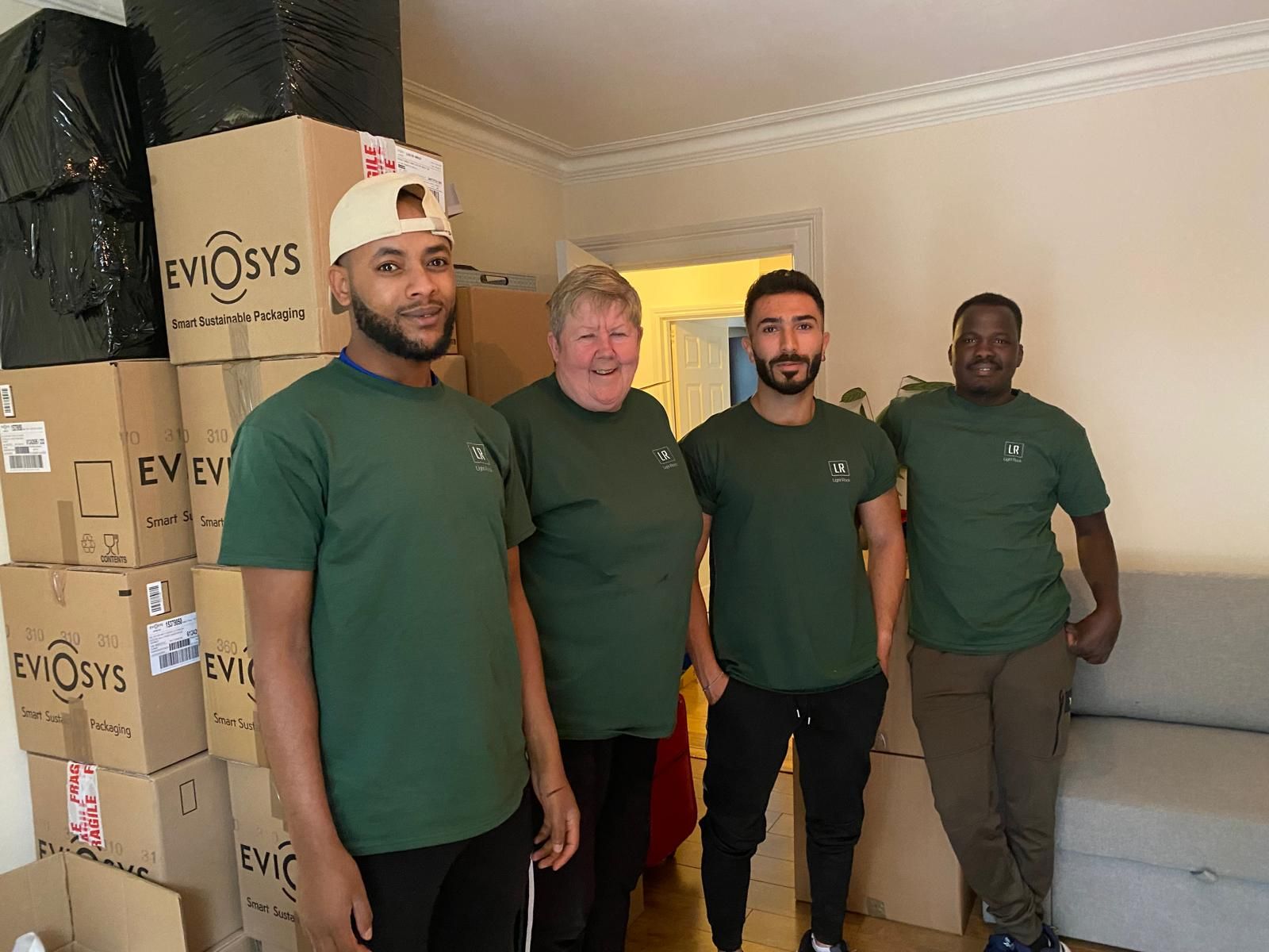 Four people, two men in green shirts and two others, stand in a room with moving boxes, possibly after a move.